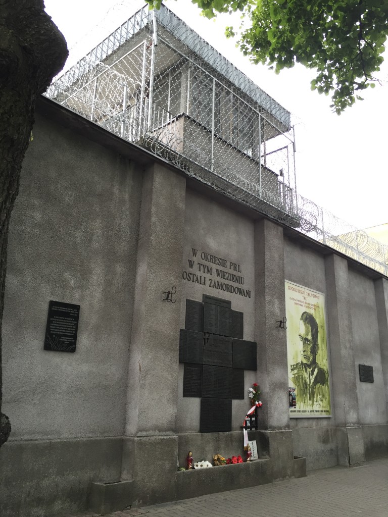 A guard tower at the Rakowiecka Prison surrounded by razor wire. Beneath is a memorial to those murdered in the prison, including Witold Pilecki and General Emil Fieldorf, pictured at the right.